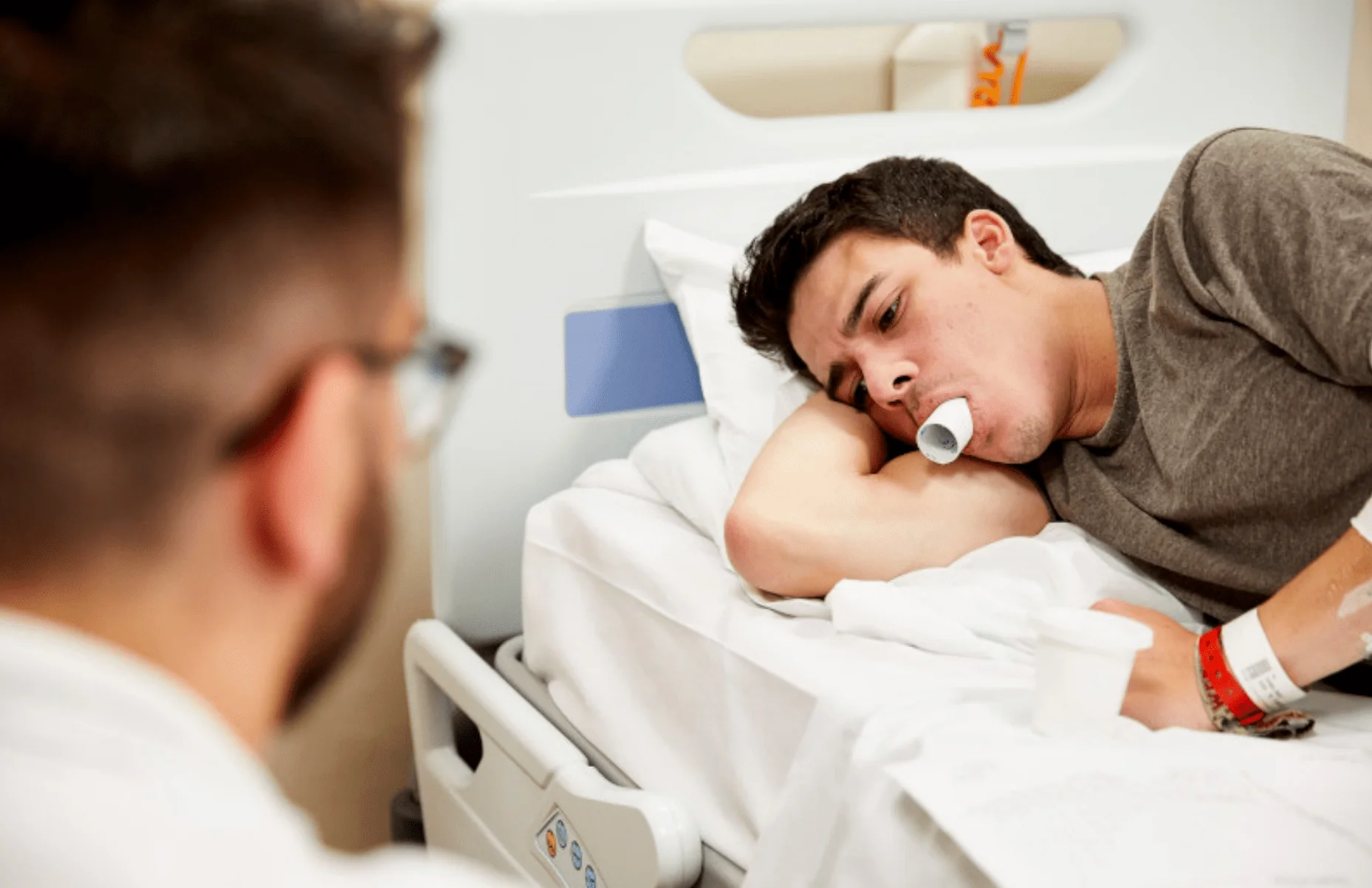 A photo of a person with Cystic Fibrosis is lying on a hospital bed with a medical device in his mouth. Another person is out of focus to the left of the frame as the camera view is over his right shoulder.