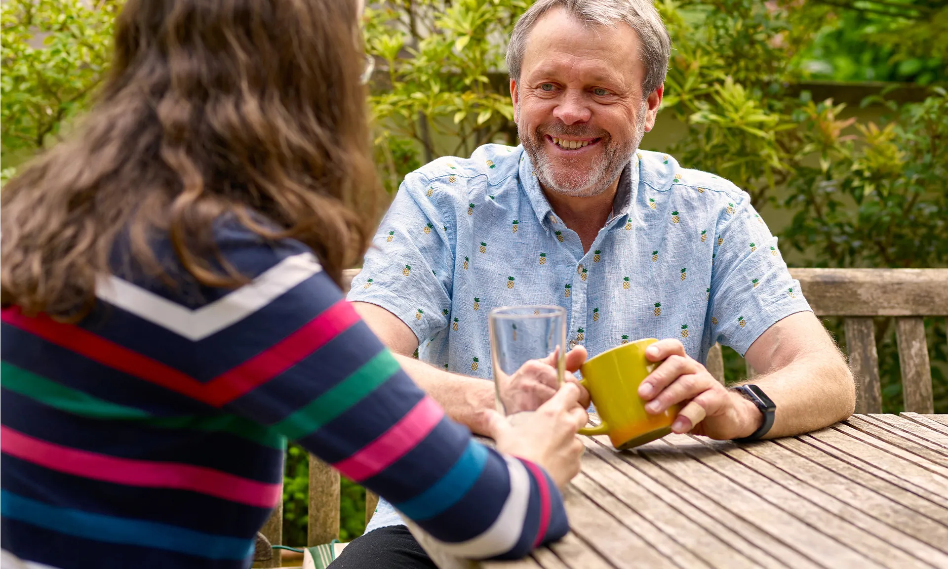 A photo of two people sitting at an outdoor table. A man in a blue shirt is sitting facing the camera is looking and smiling to his right at a woman who is positioned left of frame with her back to the camera. The man is holding a mug in both hands while the woman is holding a clear glass.