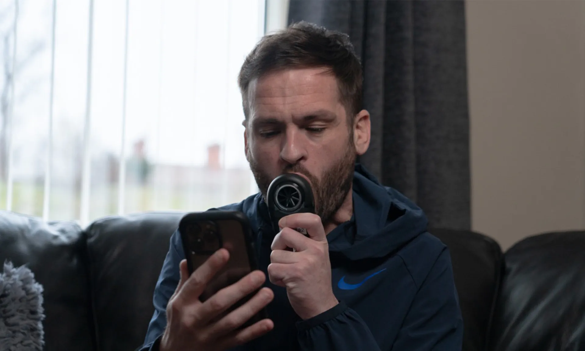 A photo of a man sitting on a sofa using a diagnostic device linked to a mobile phone. The man is centre frame and has the diagnostic device is a spirometer in his mouth with his eyes on his mobile phone towards the lower part of the image.