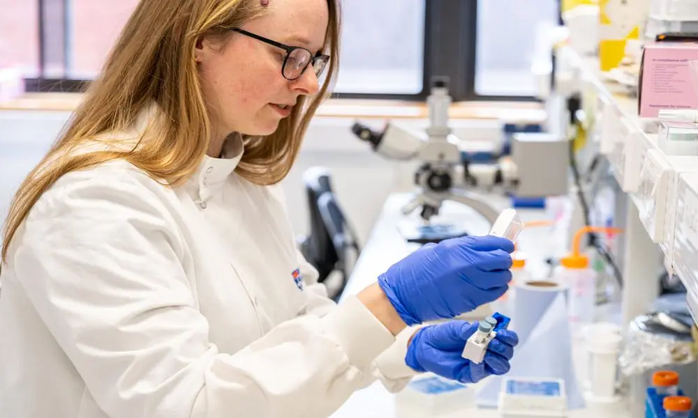 A female scientist working at a bench in a science laboratory. She's wearing a white lab coat and purple gloves.