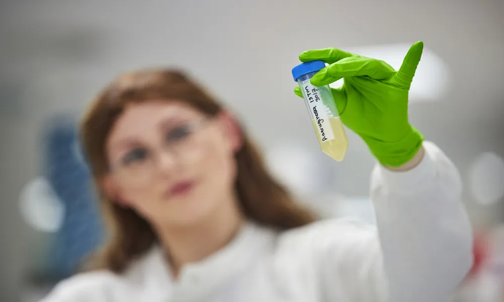 A photo of a female scientist holding a sample pot towards the camera. She's wearing a green glove and her hand and the sample pot are in focus and her face is slightly out of focus in the background of the image.