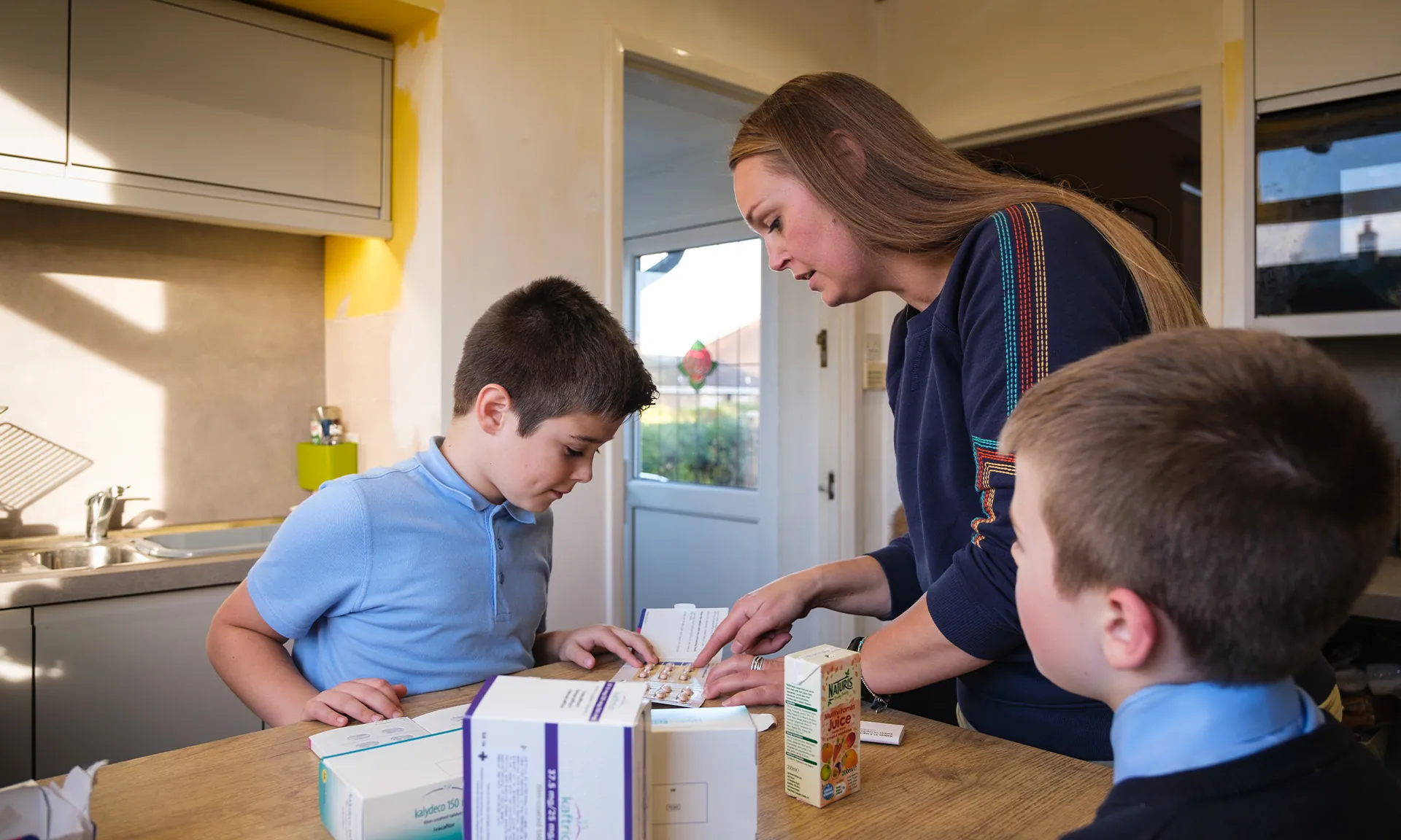 A photo of a woman and two boys. The subjects in the image are looking on a table in a room of a domestic home. The table has a collection of medicines to treat cystic fibrosis